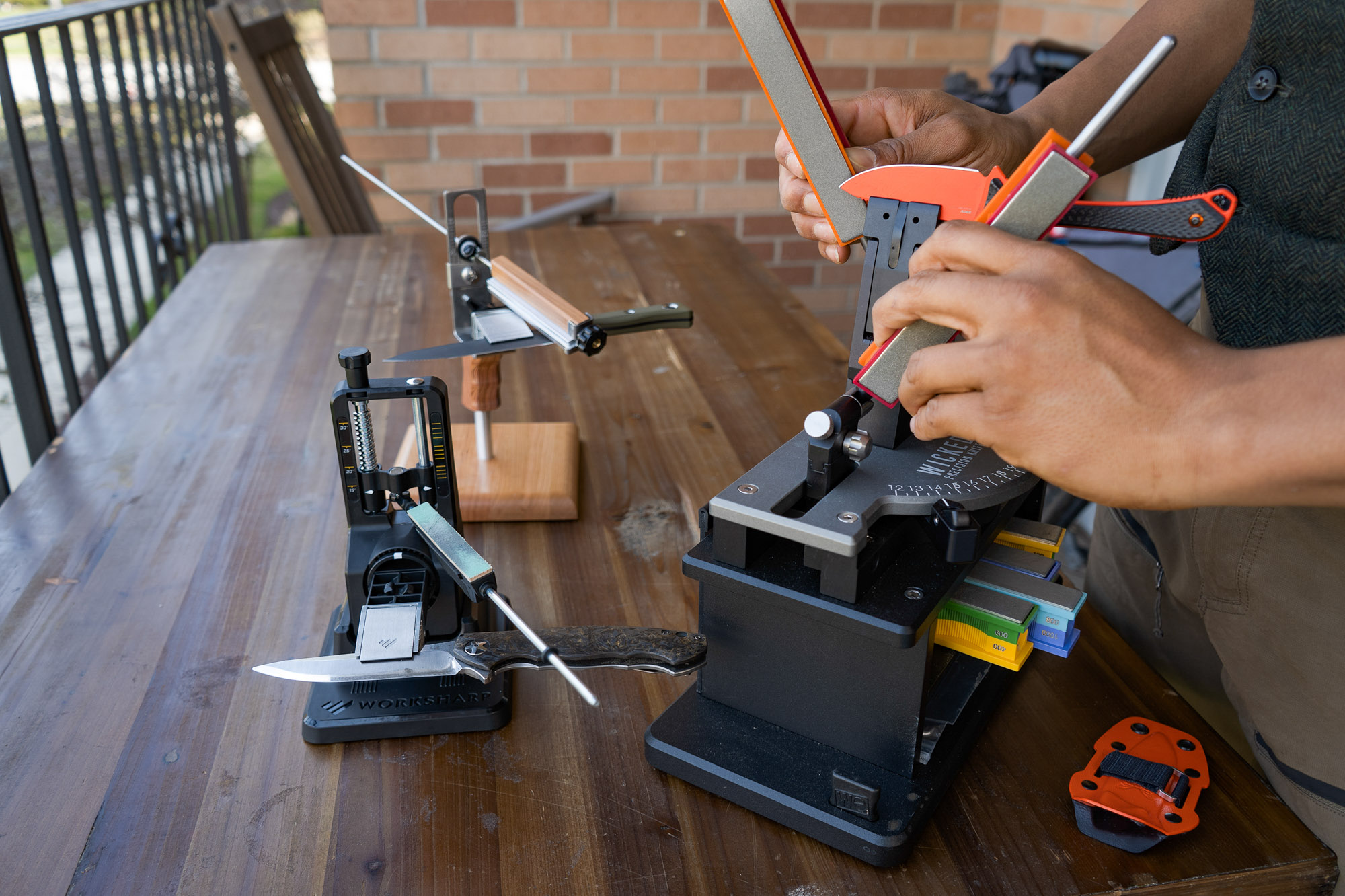 three different manual angle-guided sharpeners on a desk top, with the author sharpening a blade in a wicked edge sharpener