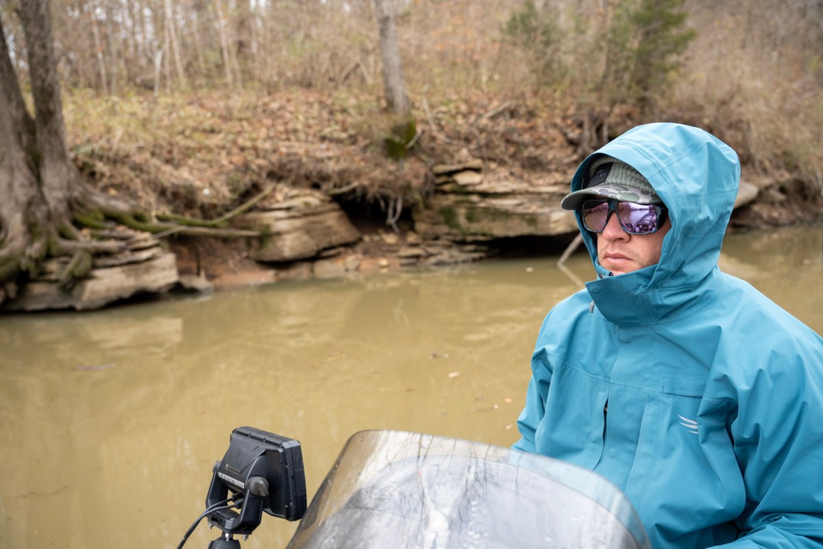 An angler driving a bass boat with full rain gear and sunglasses on.