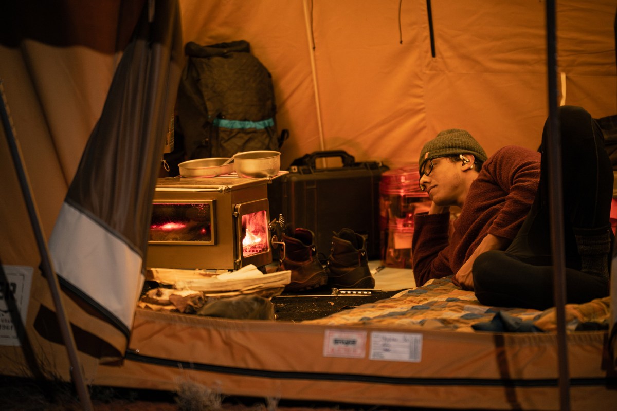 A person rests beside a glowing wood stove inside the hot tent