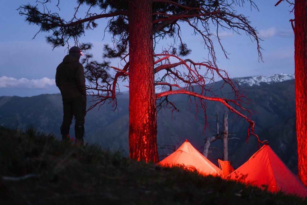 A person stands above tents lit by the headlamp’s red light in the mountains