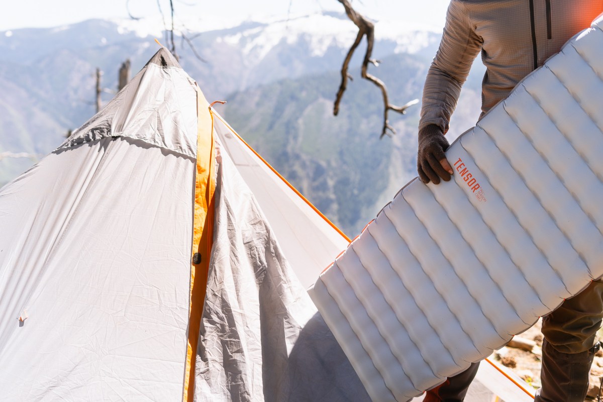 Tensor All-Season sleeping pad held beside a tent at a mountain campsite