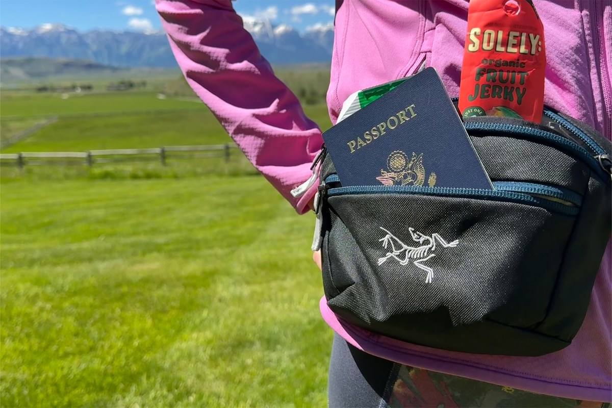 Black Arc’teryx waist pack holding a passport and snack in front of a grassy mountain view