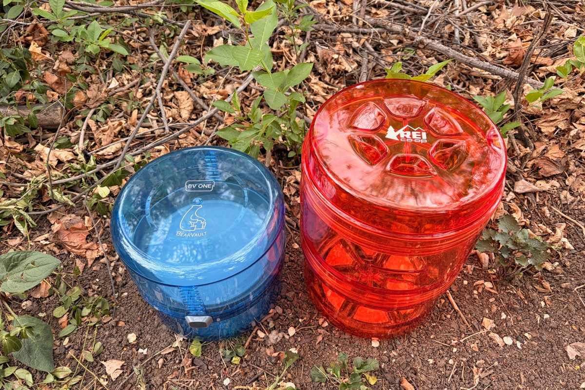 The blue BV ONE and orange REI Traverse canisters sit side by side on dirt and leaves in a wooded area