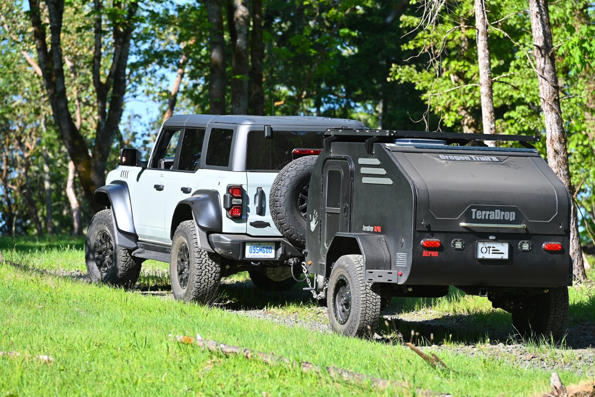 White Ford Bronco towing a black TerraDrop camper trailer through a grassy wooded area