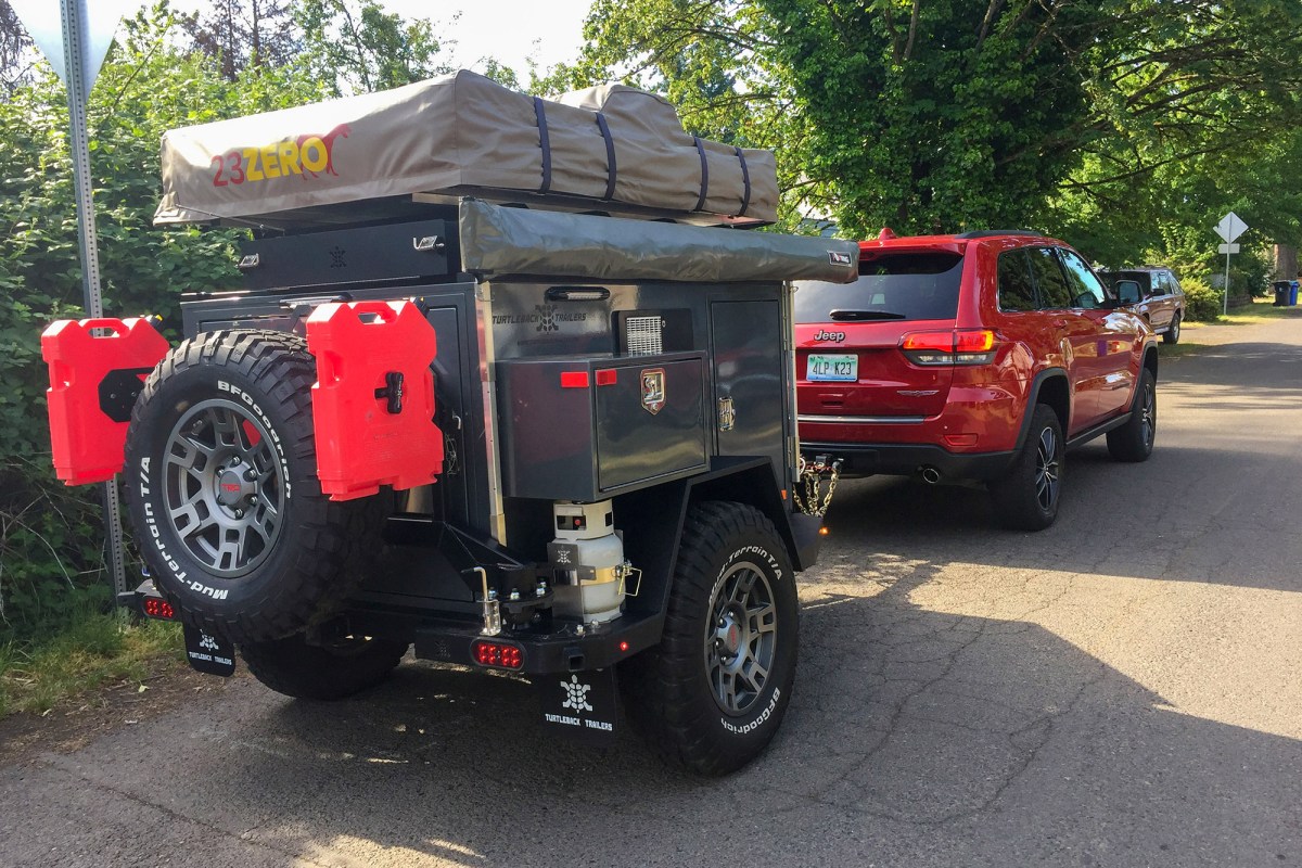Off-road camper trailer with spare tire, red fuel cans, and rooftop tent behind a red Jeep
