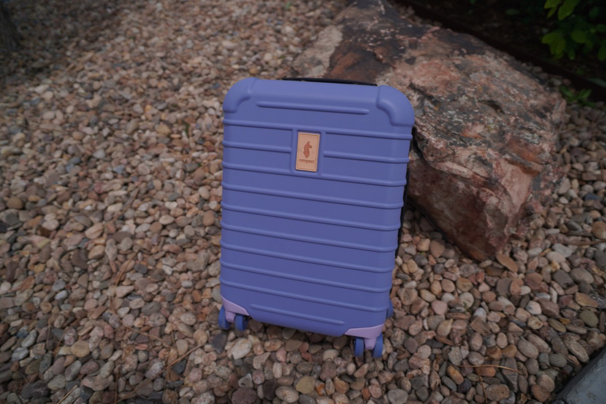 Purple Cotopaxi carry-on luggage standing on gravel beside a large rock