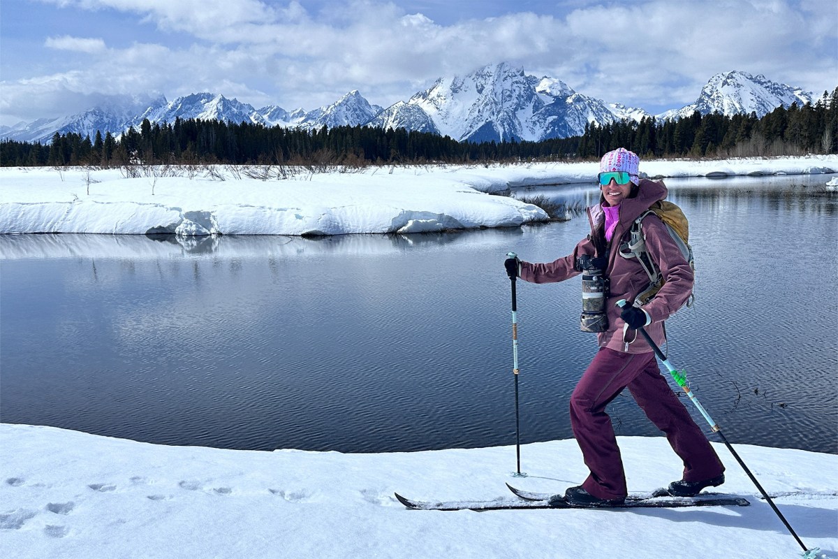 Skier in a pink fleece layer standing beside a snowy river with mountains behind her