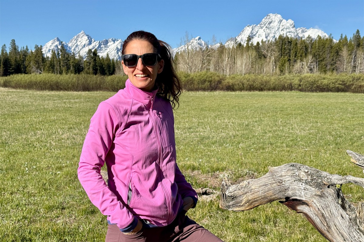 Woman wearing a pink Delta Jacket in a grassy field with mountains behind her