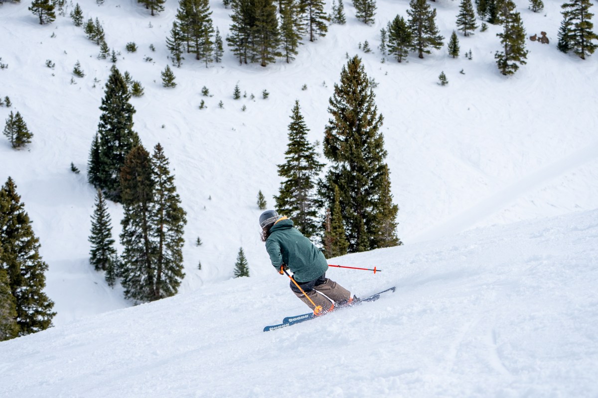 Skier wearing the Ridge Infinity Jacket 2.0 descends a snowy slope lined with pine trees