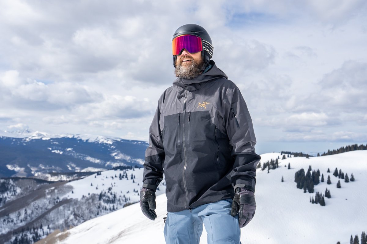 Bearded skier in Arc'teryx gray-black jacket with pink goggles, standing in snowy mountains