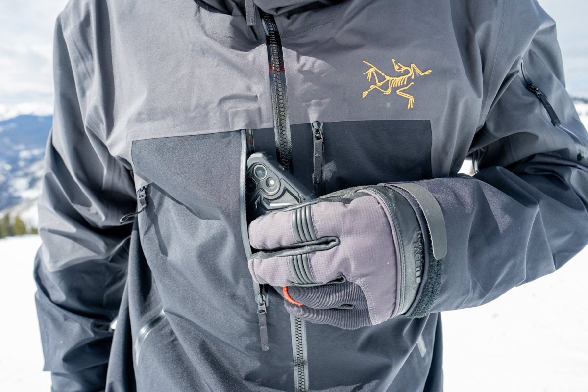 A person wearing a gray and black Arc'teryx ski jacket and gloves places a smartphone into chest pocket.