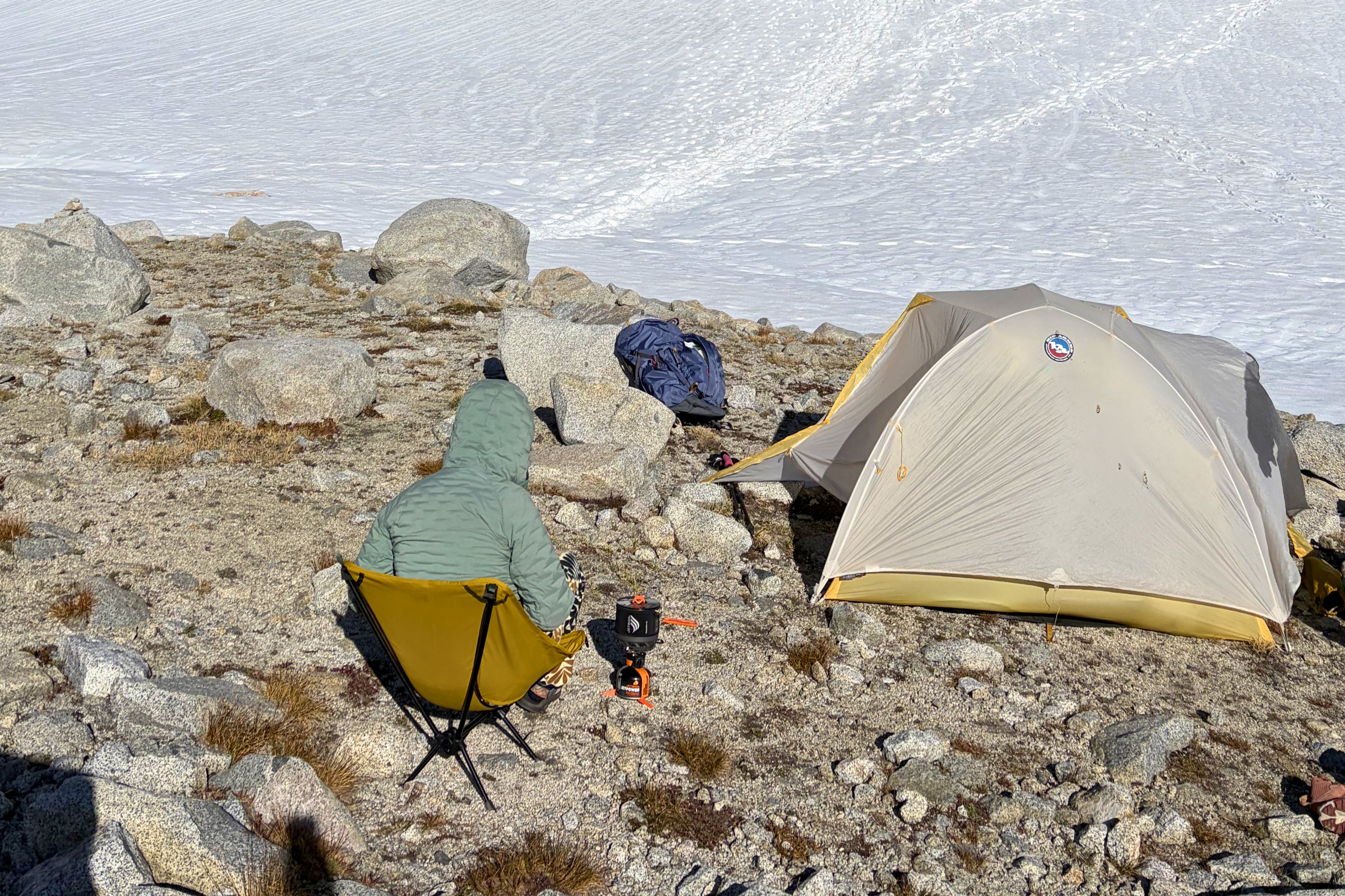 Backpacking stove cooking beside a tent and chair on rocky ground above a snowy mountain slope