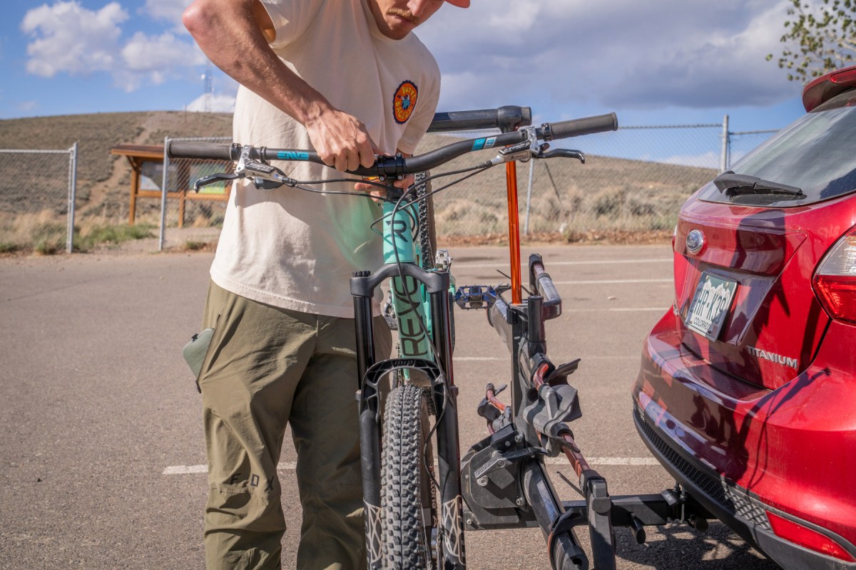 Person adjusting a mountain bike held on a hitch rack behind a red car