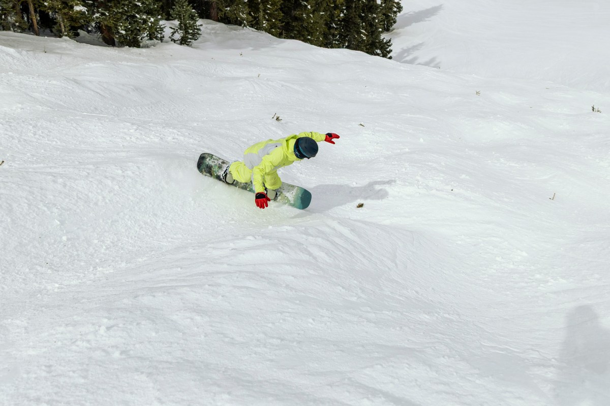 A rider leans into a turn on the Burton Custom Flying V snowboard in soft snow near trees
