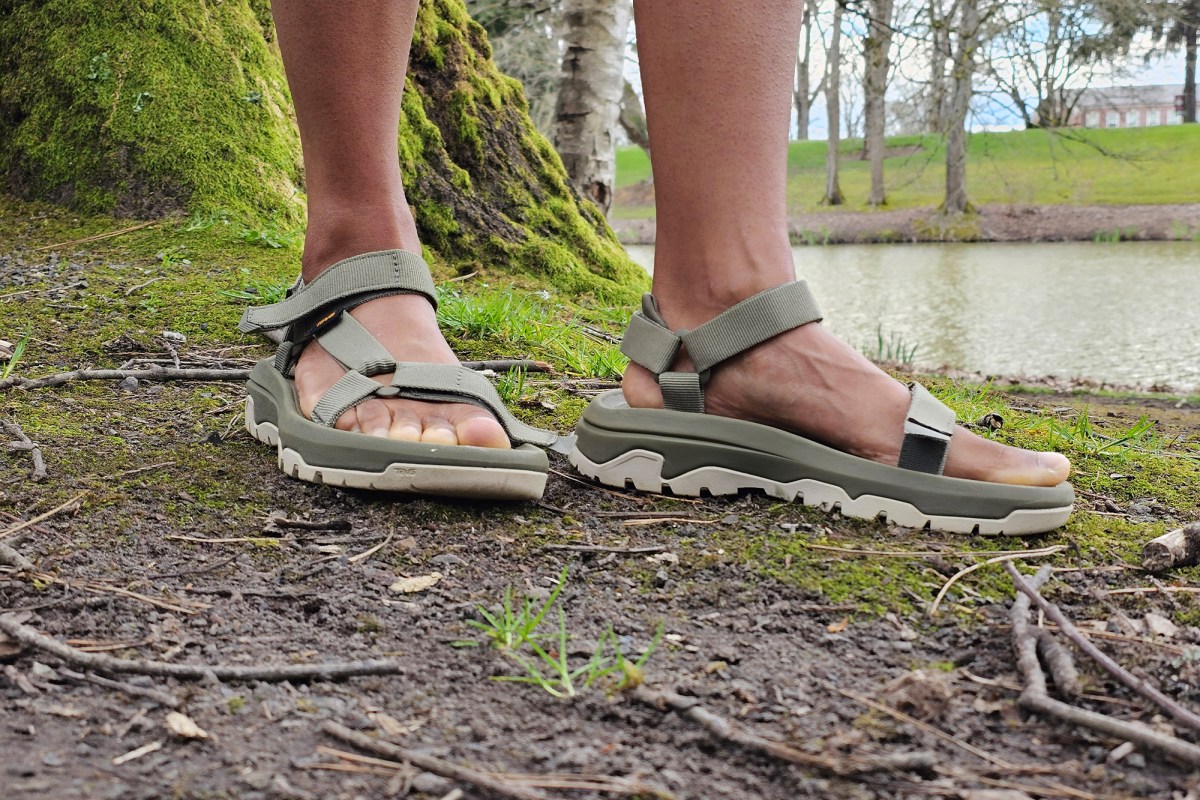 A person wearing green Teva sandals on dirt and moss near a lake
