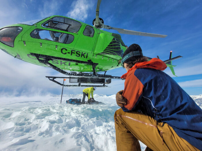 a gearjunkie tester crouches in a red and blue ortovox westalpen 3L hardshell jacket while a ski helicopter hovers above