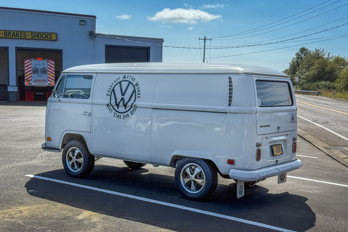 A white VW T2 panel van with a blue VW logo on the side is parked on pavement.