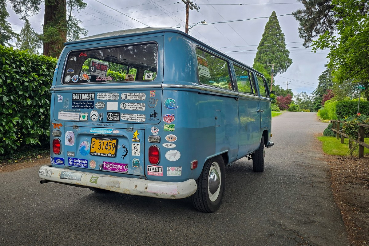 Blue vintage VW bus parked on a tree lined street with stickers covering the rear window and hatch