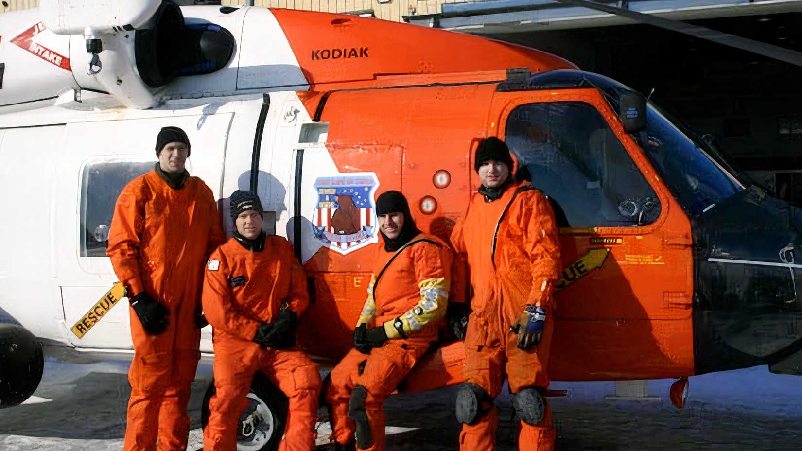 Four Coast Guard aircrew members in orange survival suits stand beside a rescue helicopter after saving 17 Alaska Ranger crew members.