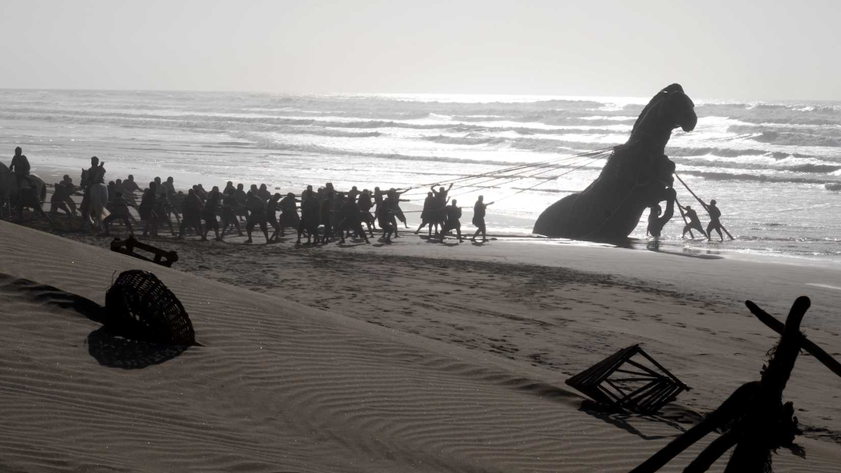 A large group of people pulls a towering wooden horse from the surf onto a beach.