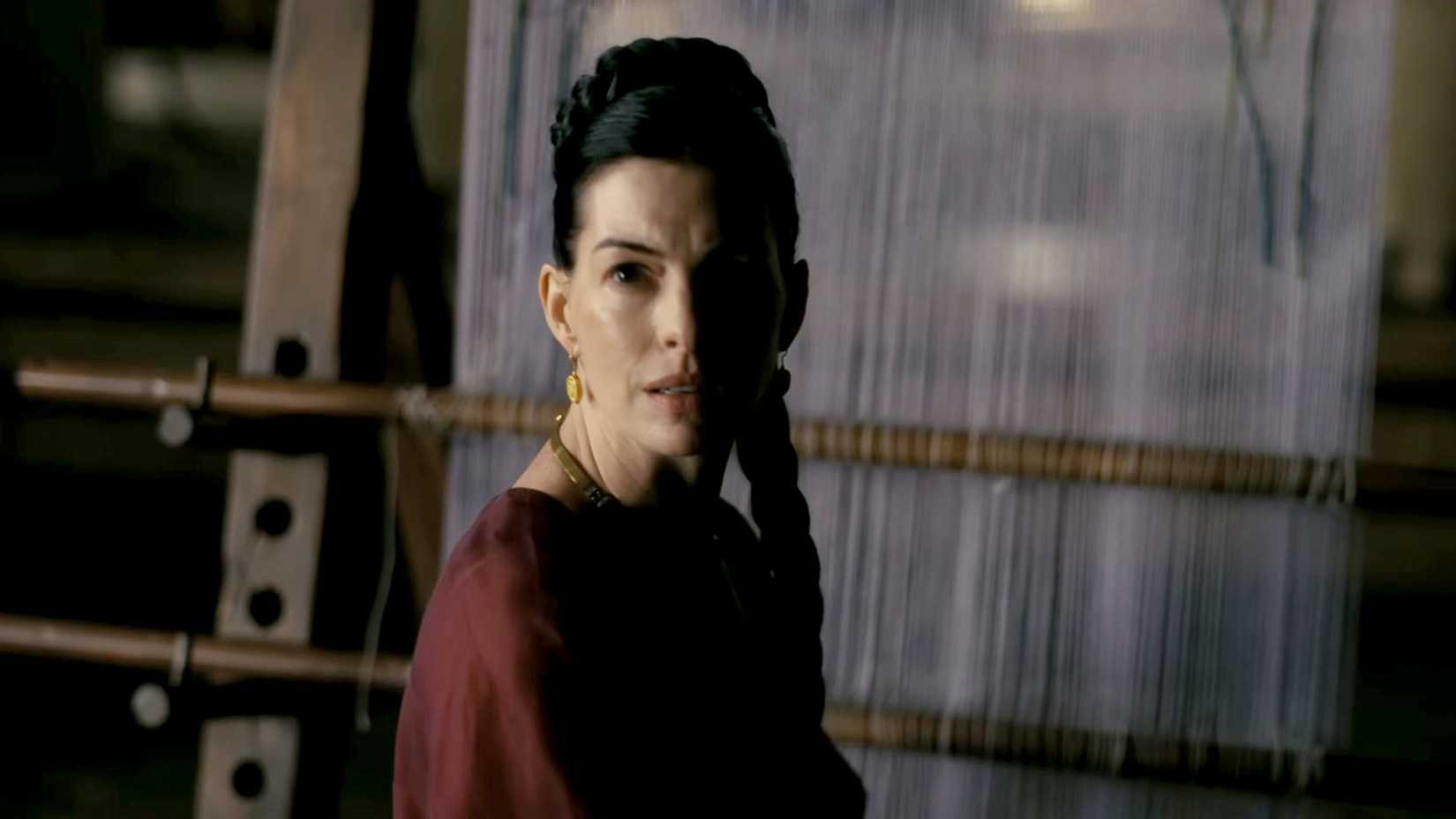 A woman in a dark red robe stands before a large wooden loom in a dimly lit palace.