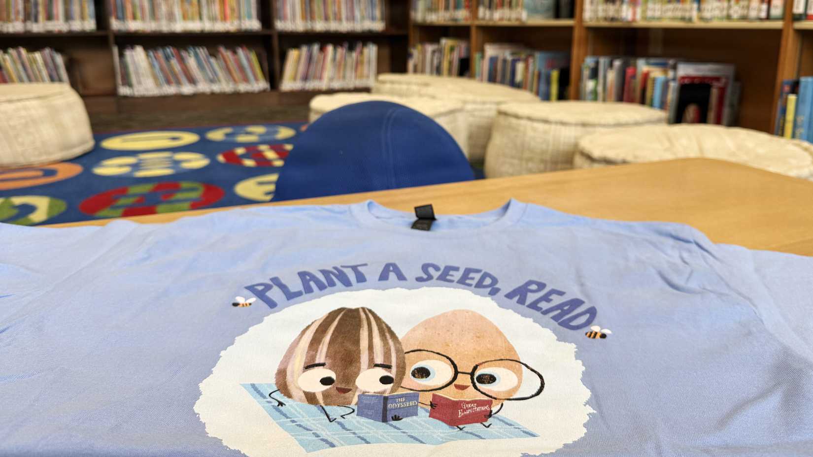 A blue “Plant a Seed, Read” T-shirt rests on a table in a military installation library surrounded by bookshelves.